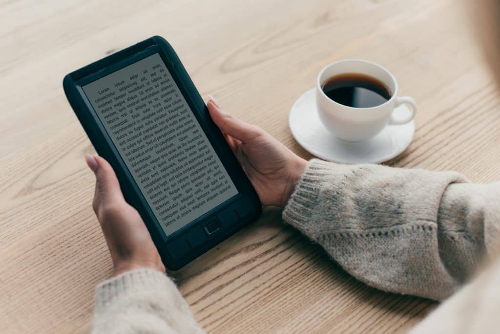 Kobiece dłonie trzymające czarny czytnik e-booków, obok filiżanka czarnej kawy