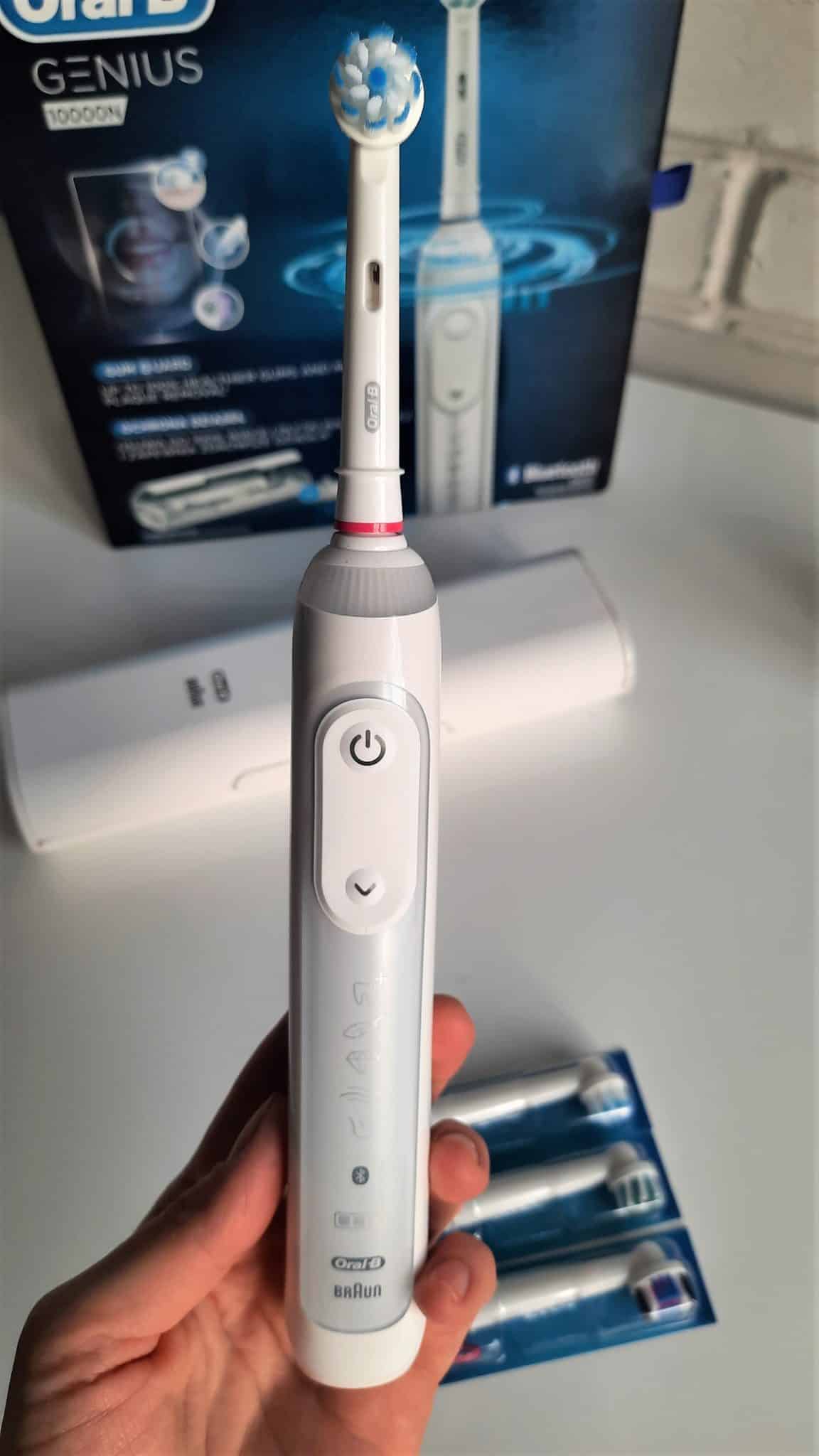 Kobieca dłoń trzymająca szczoyeczkę elektryczną Oral-B Genius 10000N, w tle pudełko szczoteczki, etui i wymienne koncówki