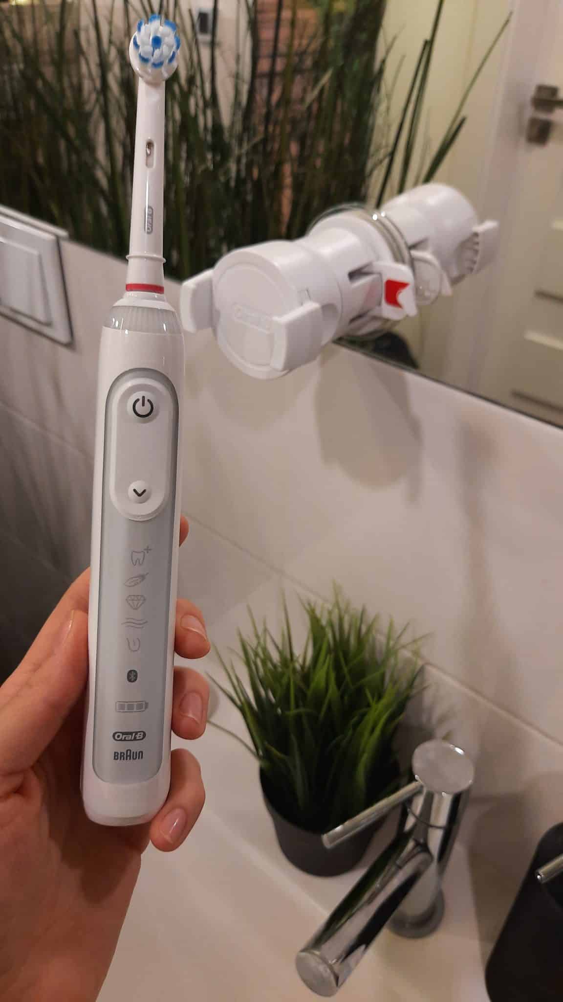 Kobieca dłoń trzyma szczoteczkę Orał-B Genius 10000N, w tle umywalka, bateria łazienkowa i lustro z uchwytem na telefon