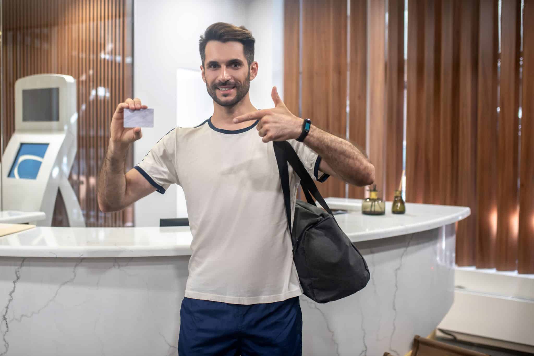 Mężczyzna ze sportową torbą trzymający w dłoni karnet na siłownię; w tle recepcja klubu fitness