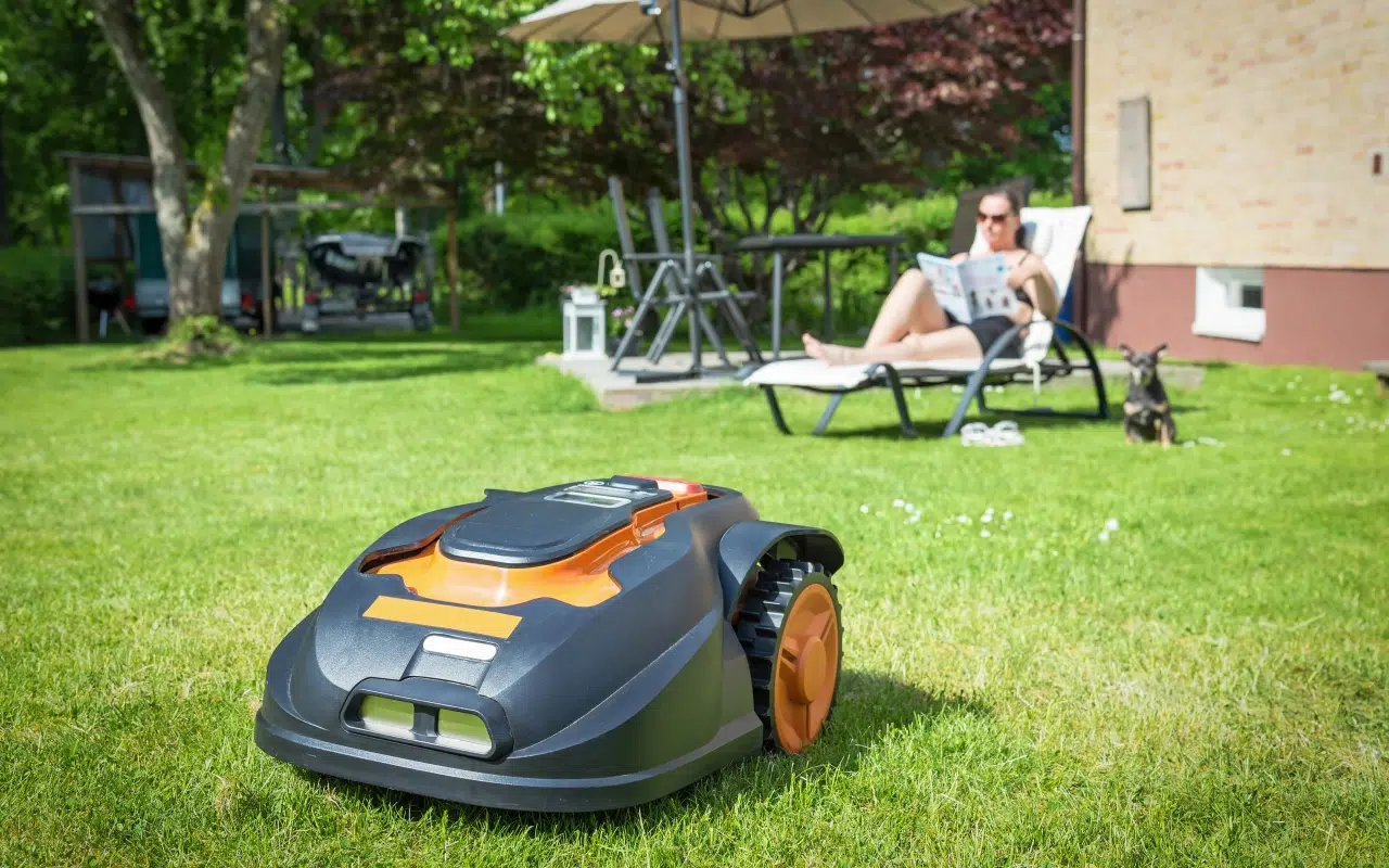 robot koszący do 500 m2 kosi trawę w ogrodzie; w tle kobieta odpoczywająca na leżaku