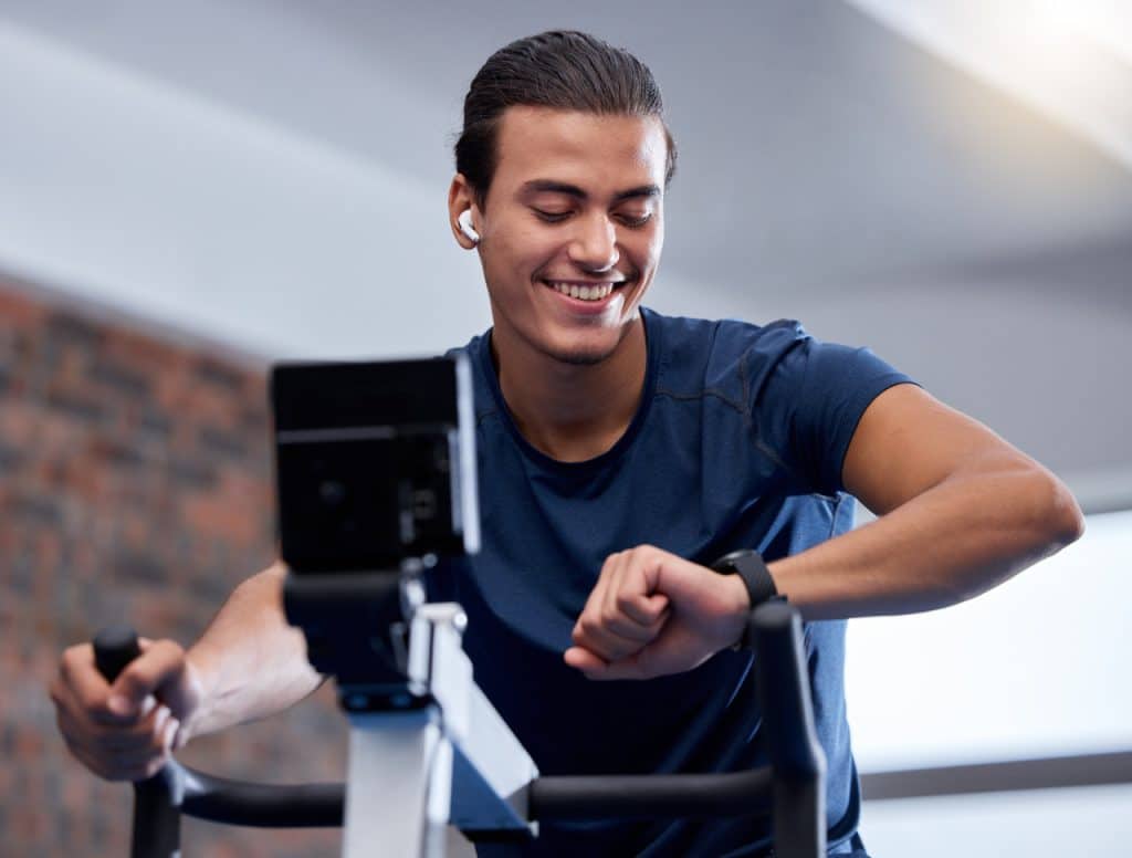 młody, aktywny mężczyzna korzystający ze sportowych gadżetów, smartwatcha, słuchawek bezprzewodowych i rowerka stacjonarnego