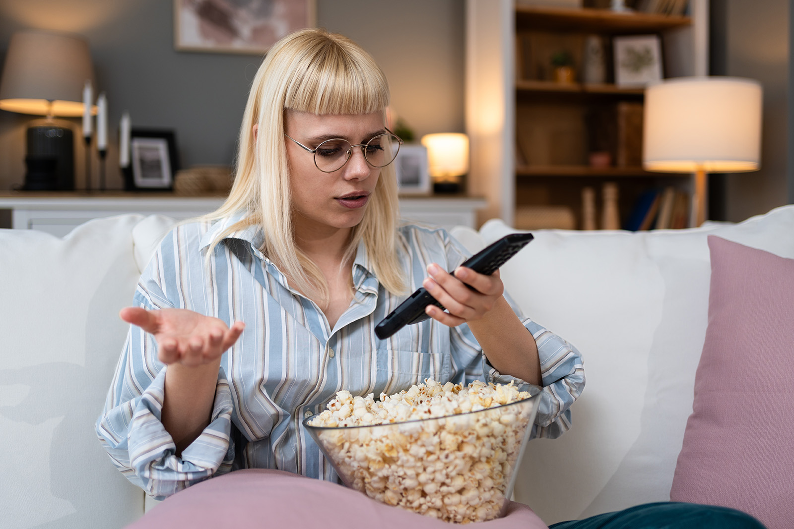 Kobieta siedzi na kanapie z miską popcornu, próbując obsługiwać telewizor pilotem. - tytułowa
